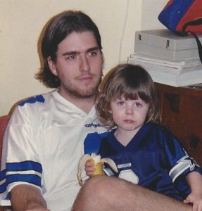 A young Hallie sits in Chris's lap while they both wear Troy Aikman jerseys